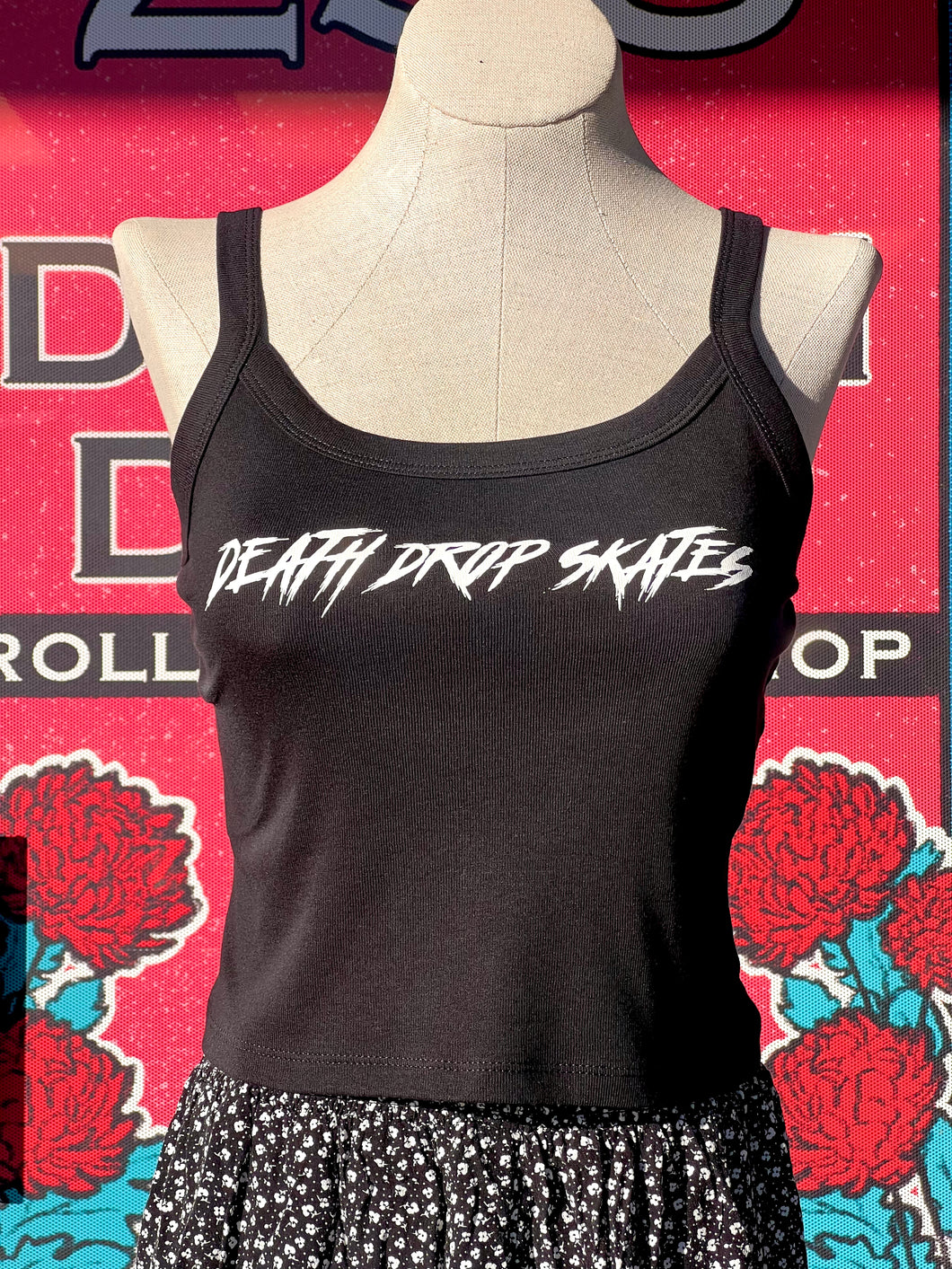 a black tank top with white lettering that says Death Drop Skates on a mannequin in front of a coral pink background with red flowers